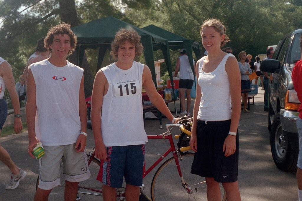 A young bike racer stands with friends and family after racing in a triathlon. He's dressed in a sleeveless white tank and blue board shorts. His unruly hair tops a broad face with a wide smile.