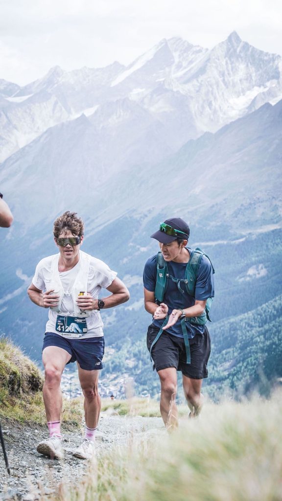 Keir Plaice passes a fellow competitor in the Matterhorn Ultraks. His hands are gripping the sides of his white ultra vest as he strides purposefully forward. The man he's passing turns to offer encouragement as he claps. A massive Alpine valley yawns behind them.