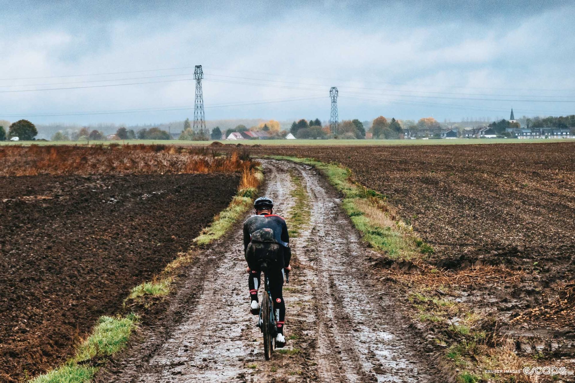 A single rider makes his way along a muddy cobbled road, between two muddy fields. 