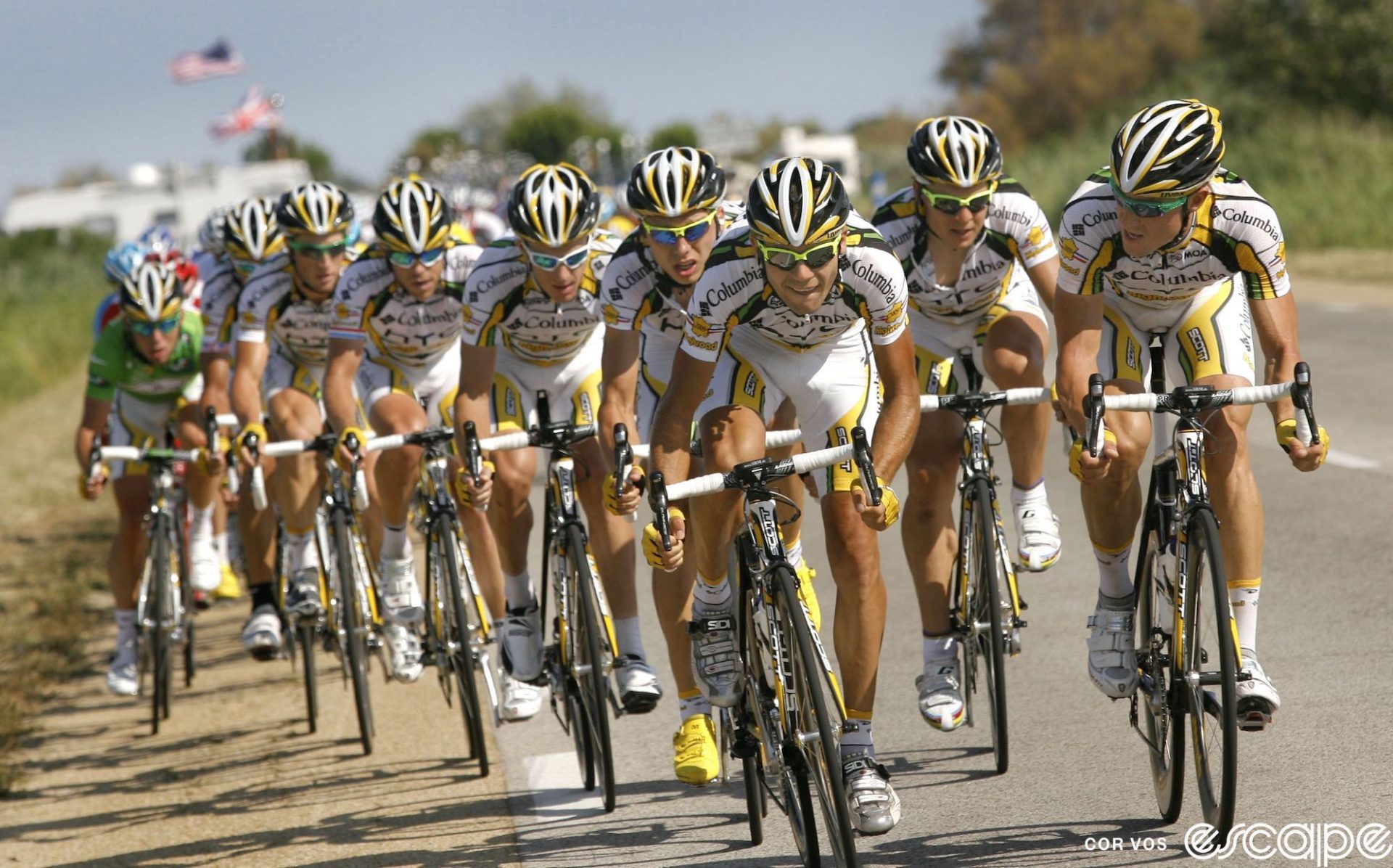 The entire HTC-Columbia team rides on the front of a race. Eight riders all in the distinctive white, yellow, and black kit ride in a double paceline. They're slammed over in the gutter and at the back a green-jersey clad Mark Cavendish rides in the dirt on the side of the road.