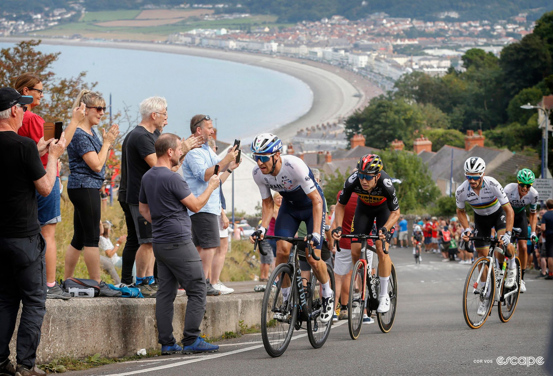 Michael Woods, Wout van Aert and Julian Alaphilippe tackle the Great Orme in 2021. The three are shown high on the climb, looking down on a curved beachfront and city behind.