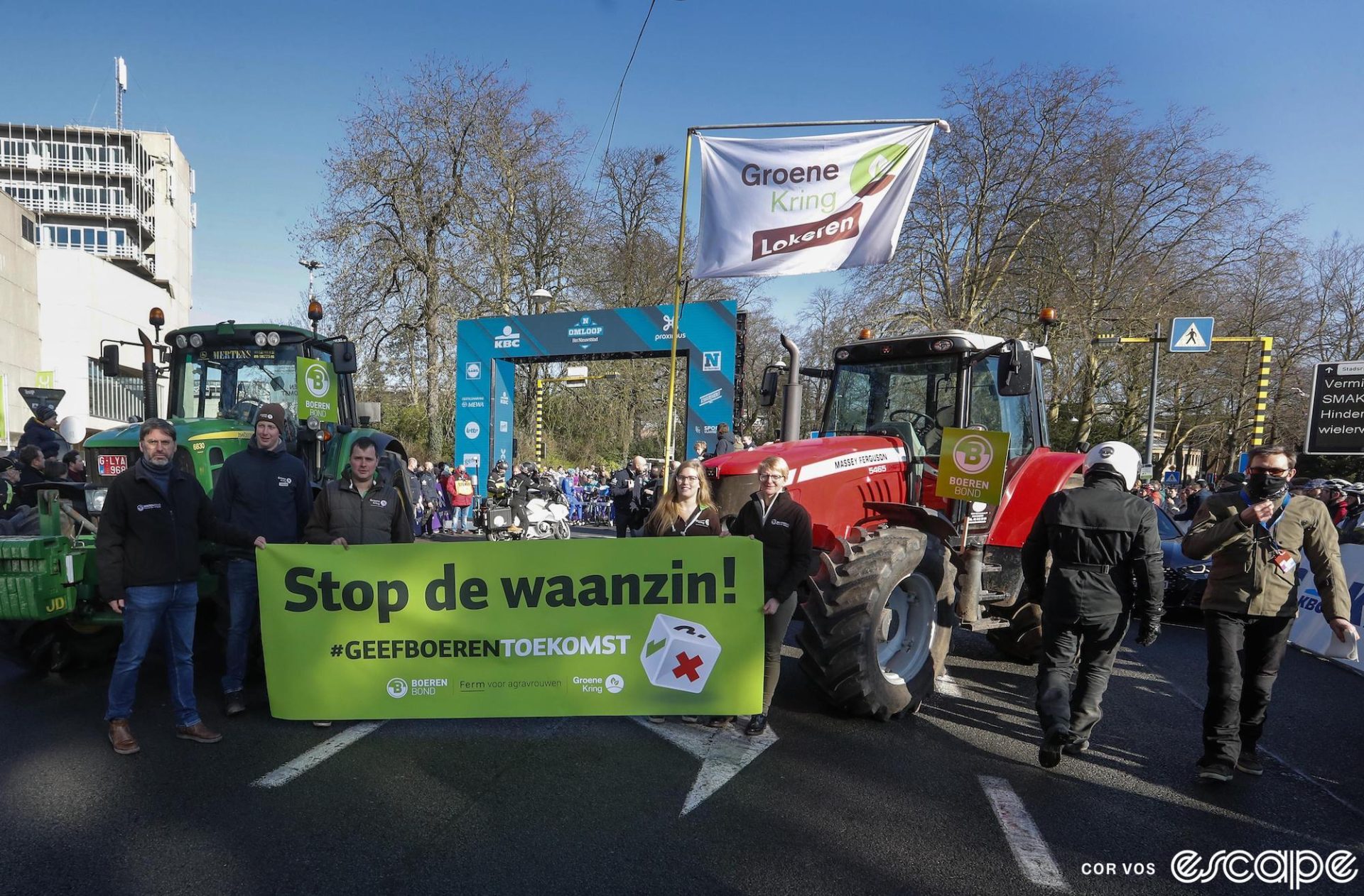 Two tractor sit parked in front of a start-line gantry for Omloop Het Nieuwsblad. Several farmers pose in front with a large green banner that reads, in Dutch, "Stop the madness! Give farmers a future."
