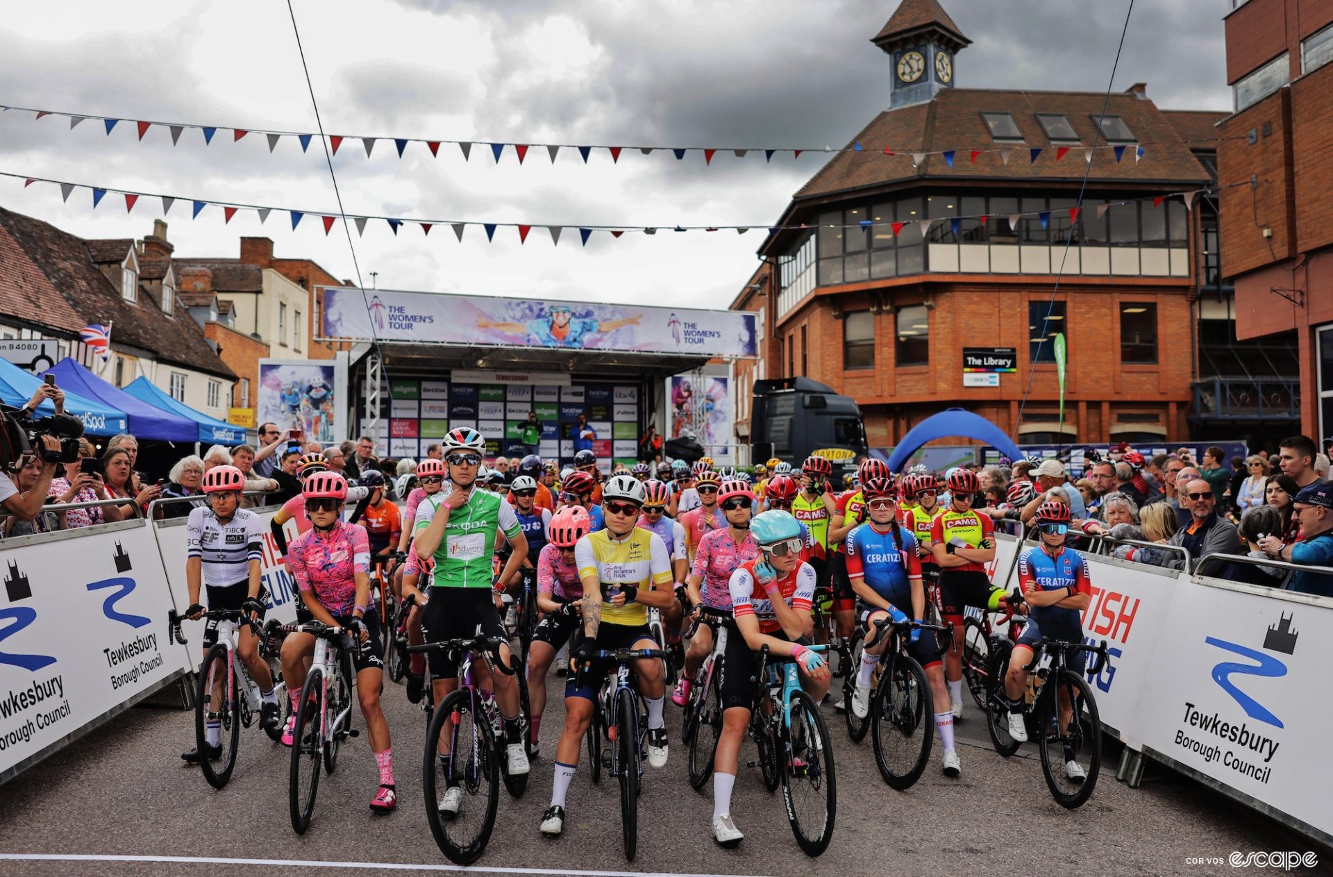 The peloton awaits the departure of stage 3 of The Women's Tour in Tewkesbury. The pack is standing on their bikes ready to go, with fans clustered at the barriers. The now-empty sign-in stage rises behind them.