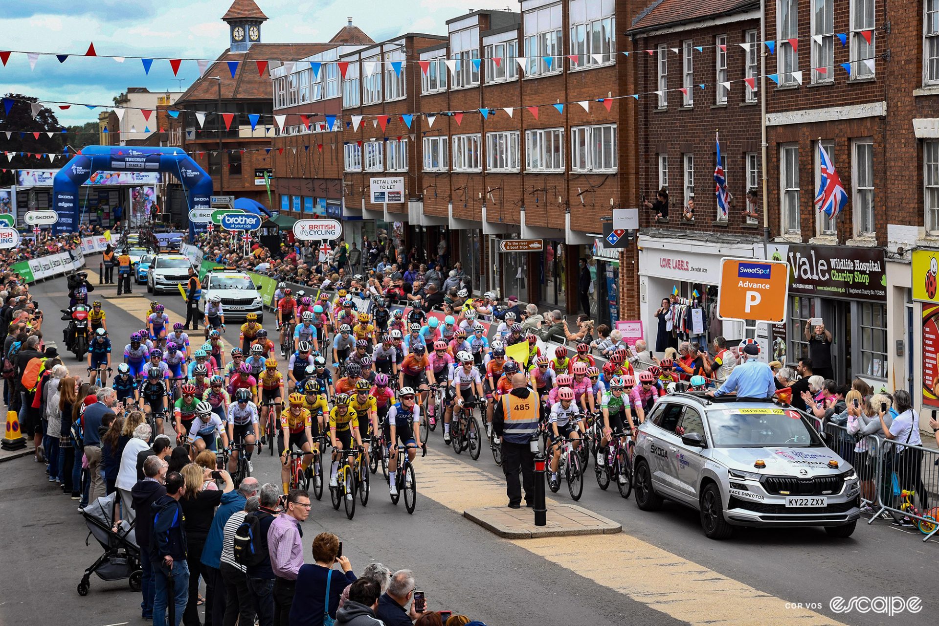 The 2022 Women's Tour starts in Tewkesbury on stage 3. The pack roll out behind the race director's car in a downtown area with fans lining both sides of the road.