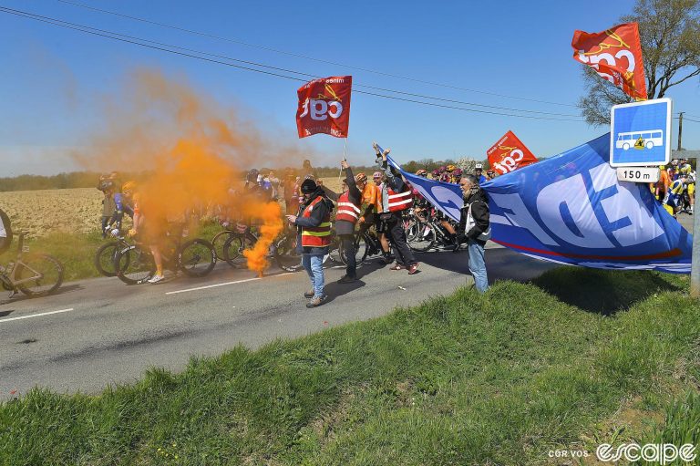 Bike racers pass through an orange cloud of smoke from a road side flare set off by protesters. The ride on the far side of the road as security attempts to corral the protesters and their banner to the side.