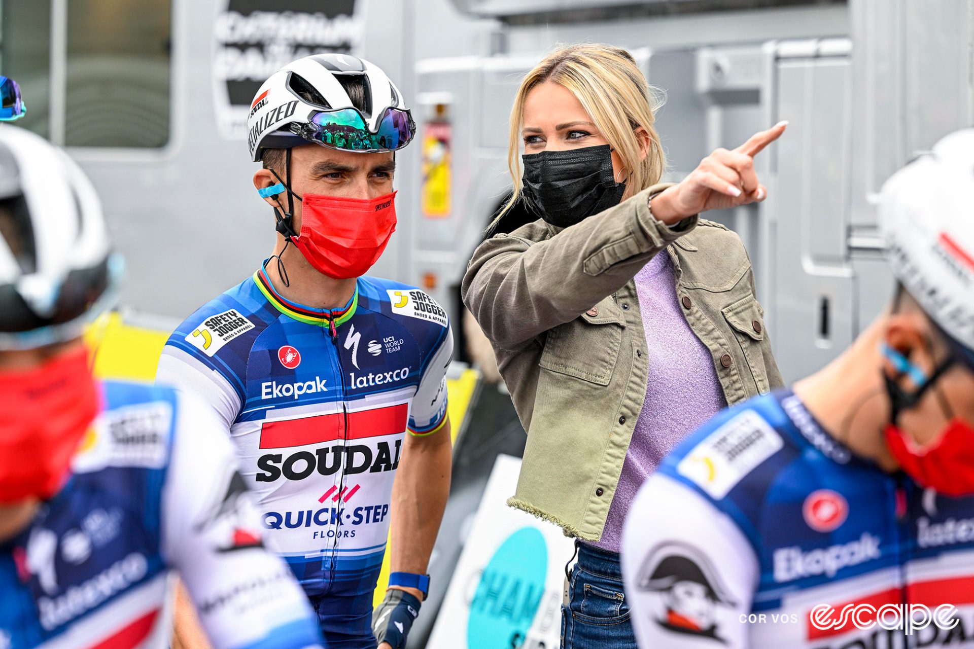 Julian Alaphilippe and Marion Rousse at the start of a race; both are wearing masks and Rousse is facing Alaphilippe while pointing to something off-camera.