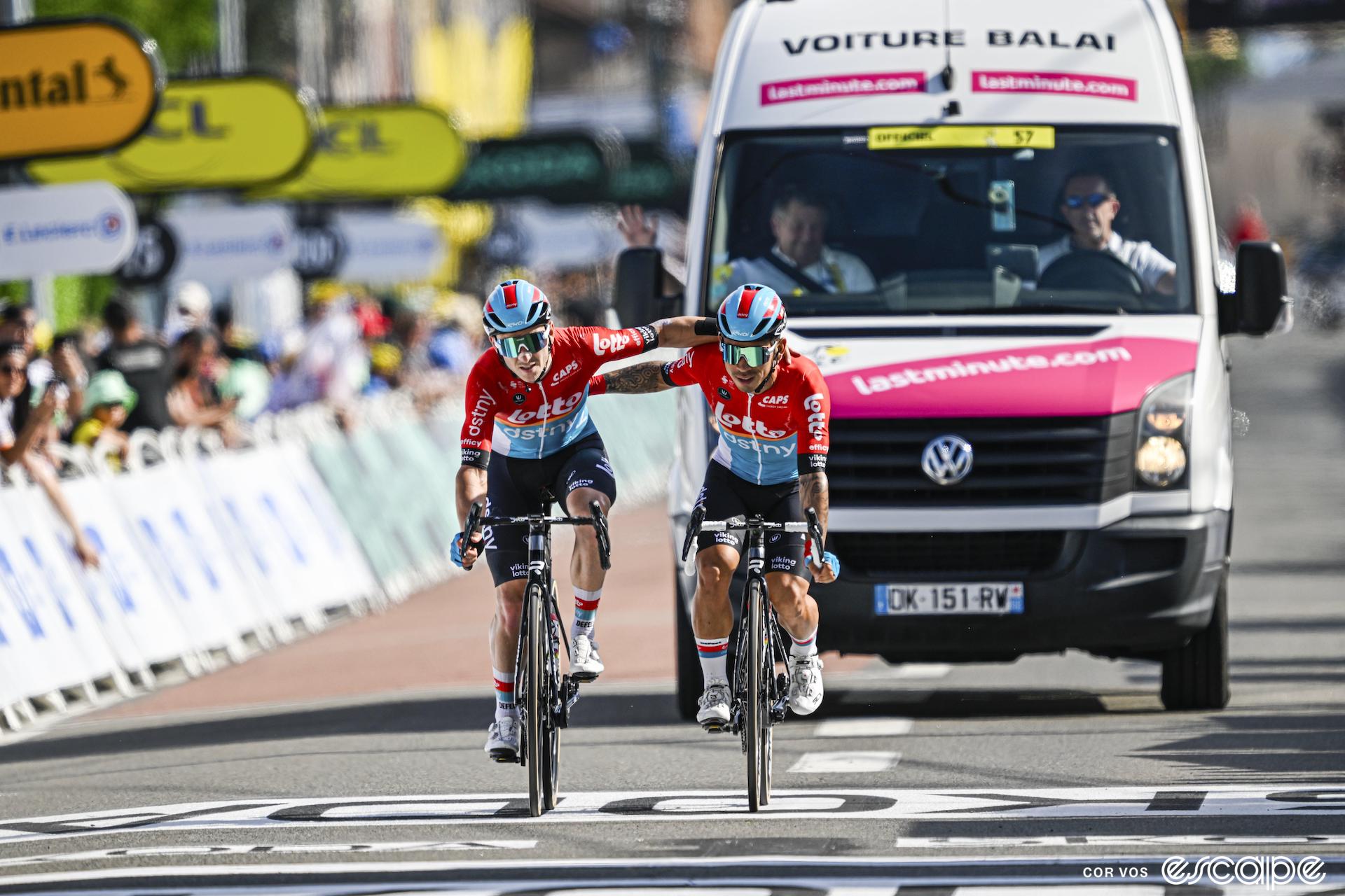 Caleb Ewan and Jasper De Buyst cross the finish line at the Tour de France, arms around each other, with the 'broom wagon' right behind them.