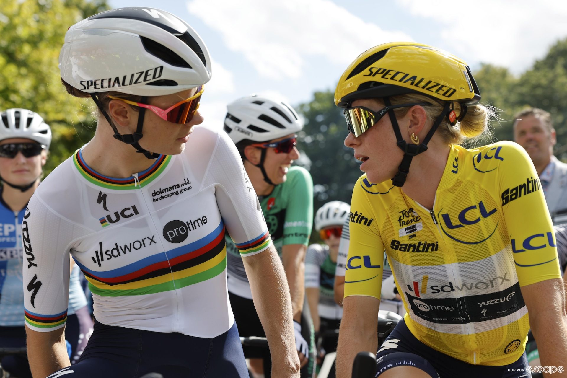 Lotte Kopecky and Demi Vollering lean in close to talk while waiting for a bike race to start. 