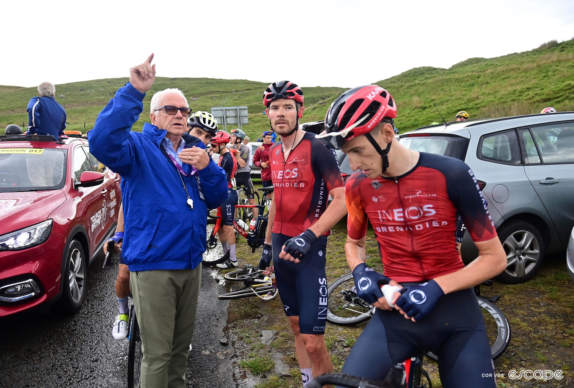 Race promoter Mick Bennett with Luke Rowe and Ben Turner at the 2023 Tour of Britain. They're standing on the side of the road in open countryside, likely after a crash or other event has temporarily neutralized the race.
