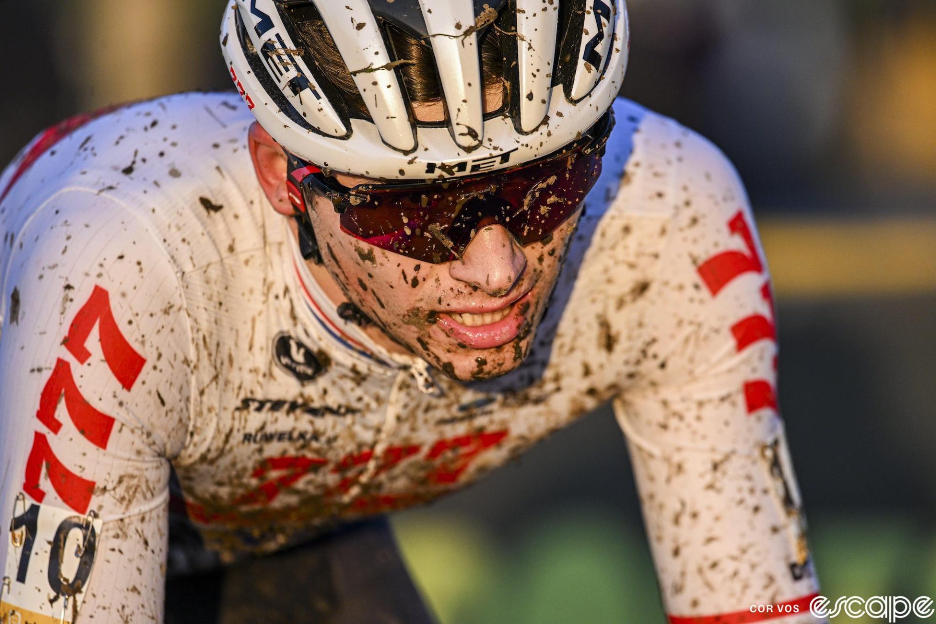 Cameron Mason races at the Superprestige round at Boom in 2023. He's shown close up, his face and white British champion's kit splattered in mud. He's wearing wraparound shades and has an intense look on his face and is lit in soft afternoon glow.