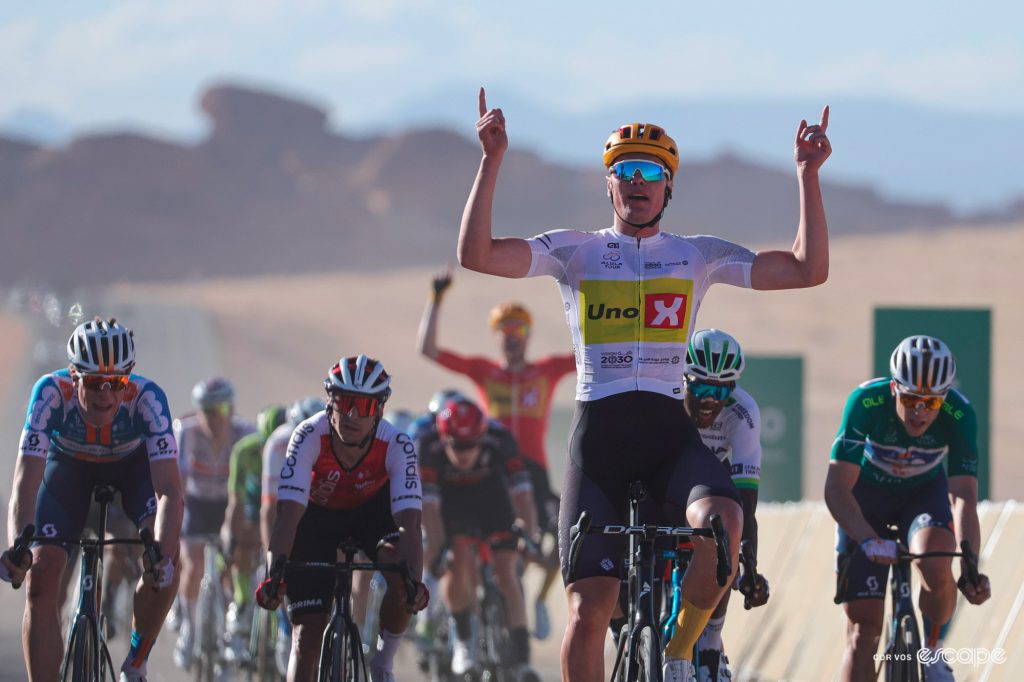 Soren Wærenskjold sits up and points both hands at the sky as he crosses the line to celebrate a stage win. Behind him, a teammate can be seen in the distance doing the same.