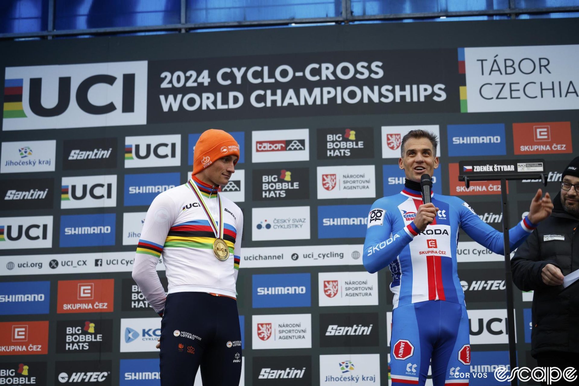 Zdenek Stybar holds a microphone as he addresses the crowd at the 2024 World Cyclocross Championships. He is standing on the podium, while next to him, winner and six-time World Champion Mathieu van der Poel looks on with respect.