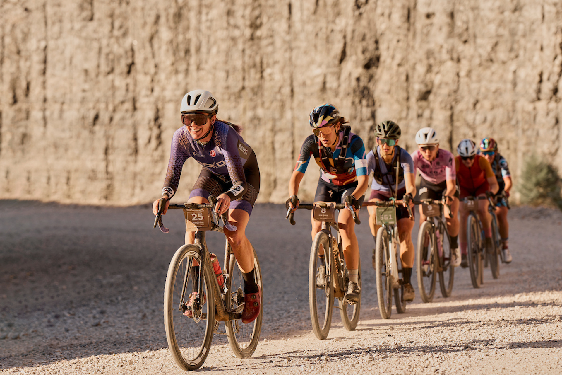 The women's field races at Big Sugar; A line of riders is stretched out on a wide gravel road with stark, steep cliff bands behind them.