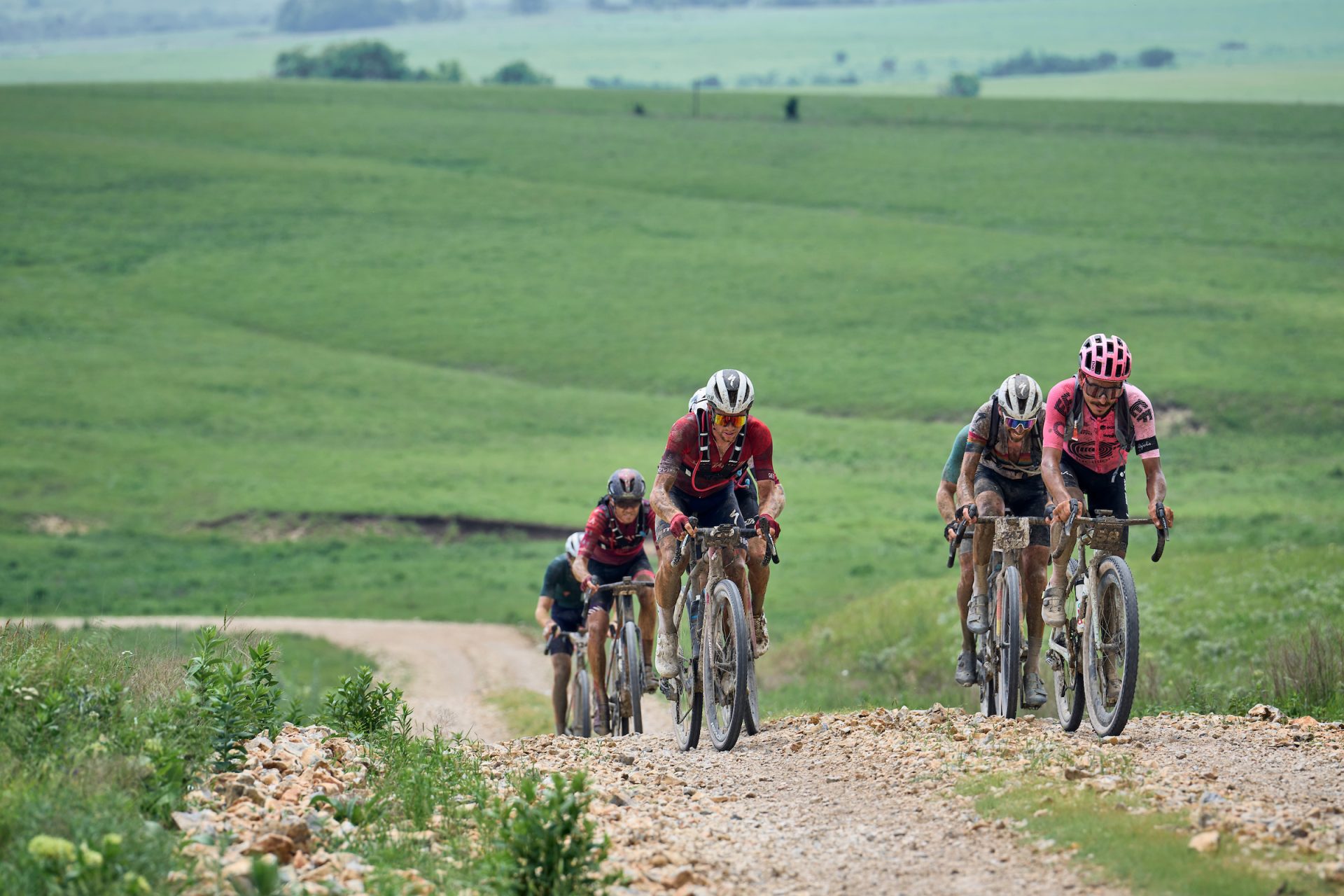 Racers at Unbound crest one of the Flint Hills' rolling rises. They're in two lines on a rocky double-track road. The riders all wear backpacks and are covered in grit and dust from the ride.
