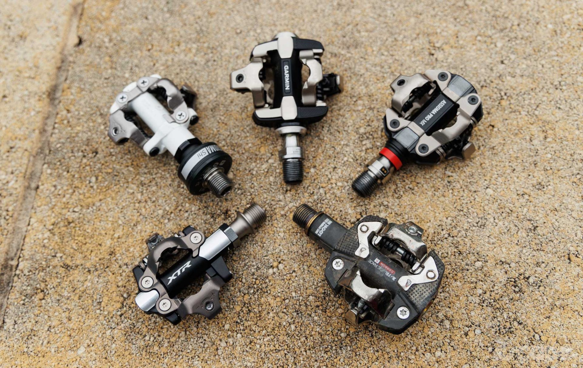 Five different SPD pedals laid out. Top right is the Favero Assioma Pro MX-2 pedals. Following in a clockwise direction, there is the Look X-Track Race, Shimano XTR, Shimano M520 with Favero Assioma Shi, and Garmin Rally XC200. 