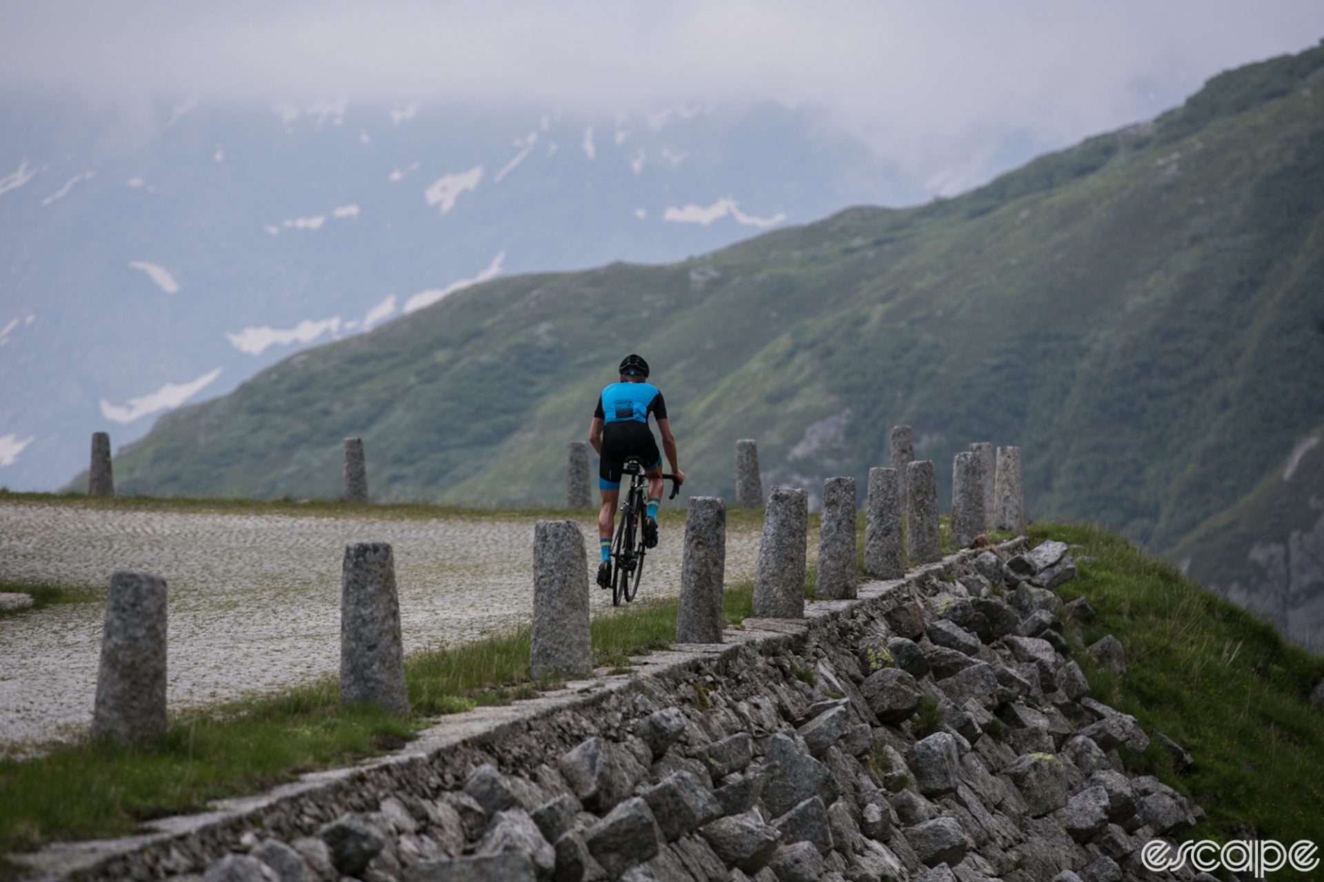 Mike Cotty rides the Gotthard Pass in Switzerland. He's riding away from the camera at a distance, nearing a switchback. The surface is paved in cobblestones and conical stone bollards frame the roadside above a steep drop. Misty mountains loom in the background.