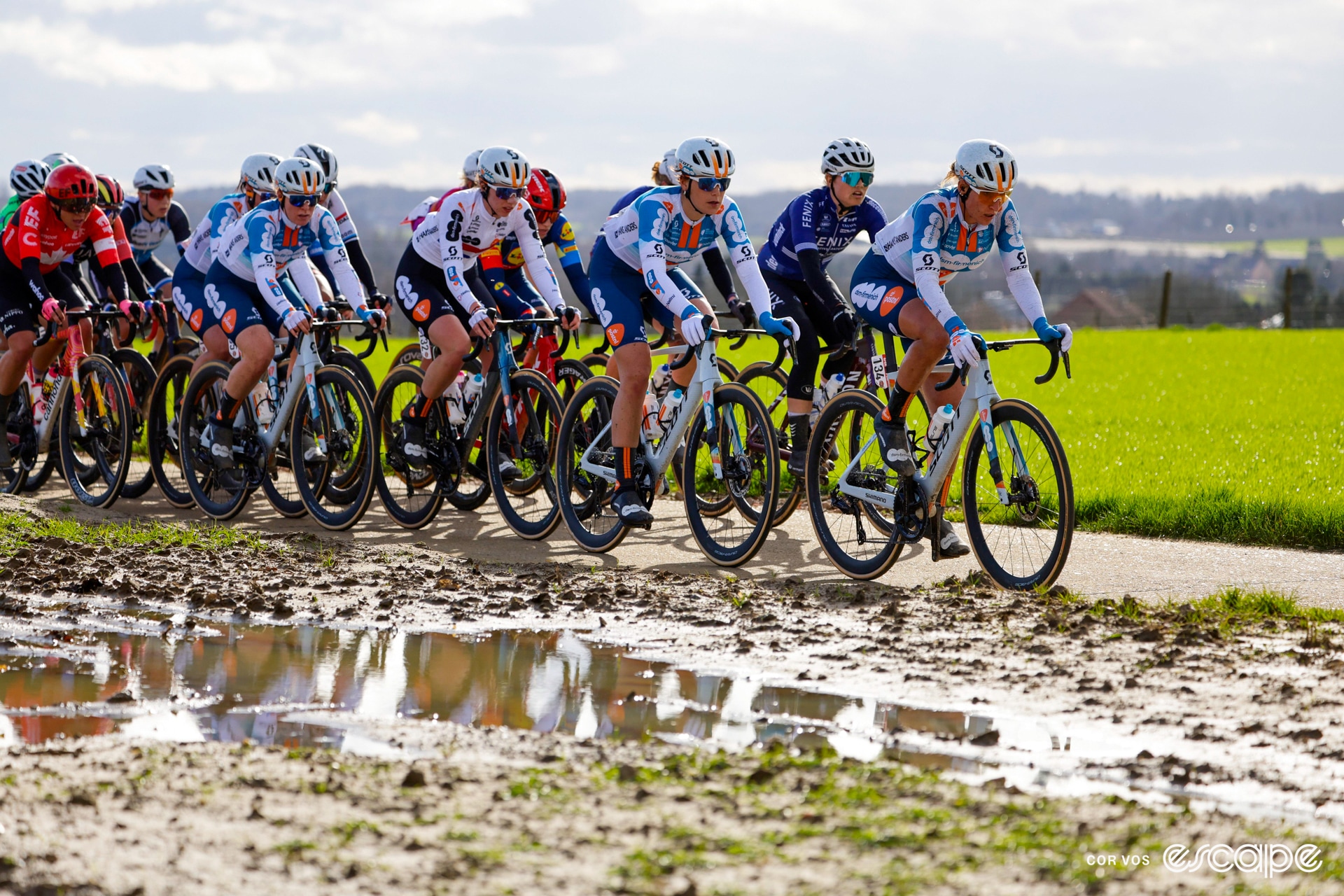 A peloton rides along in front of some mud
