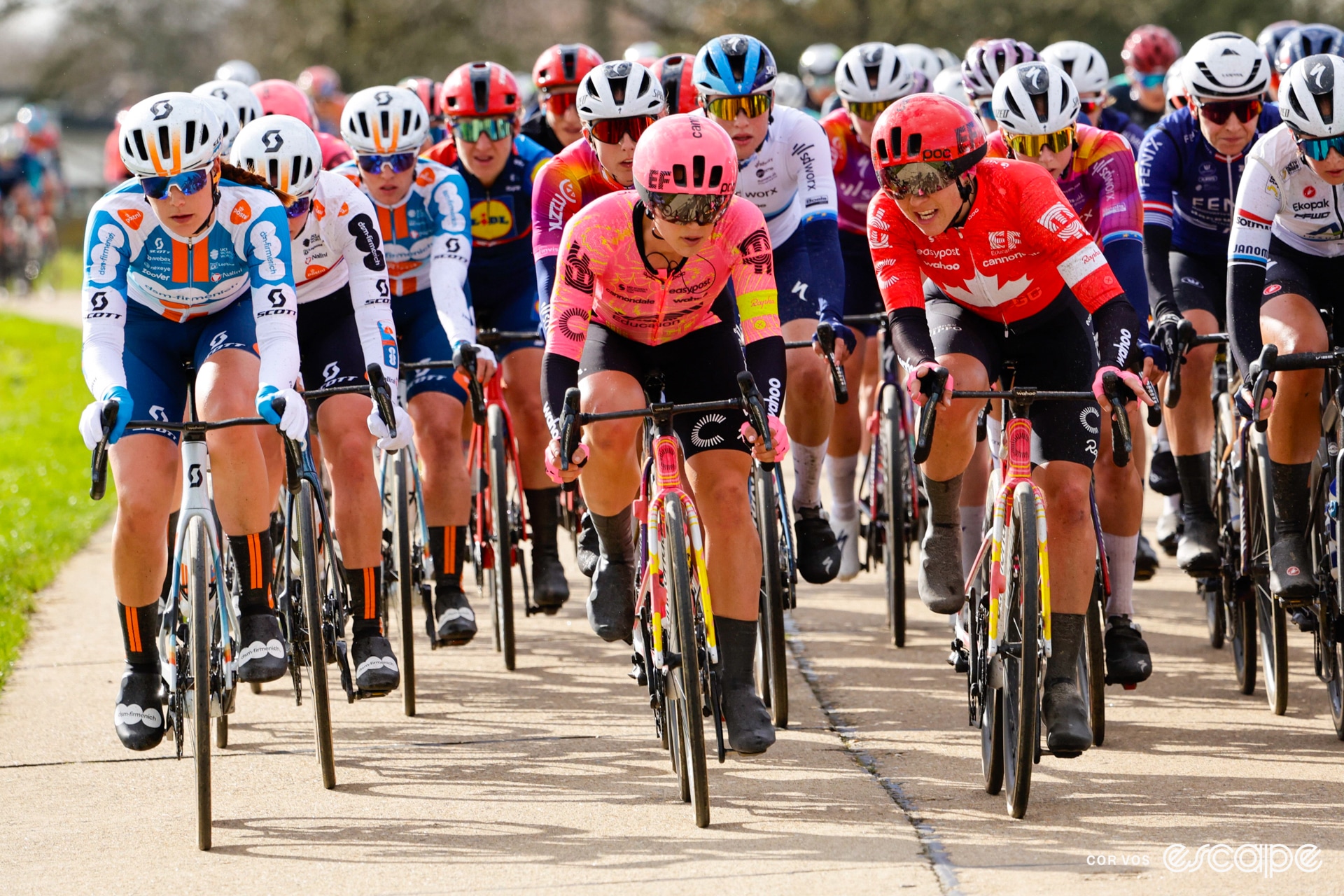 A peloton rides along, with two riders talking at the front.