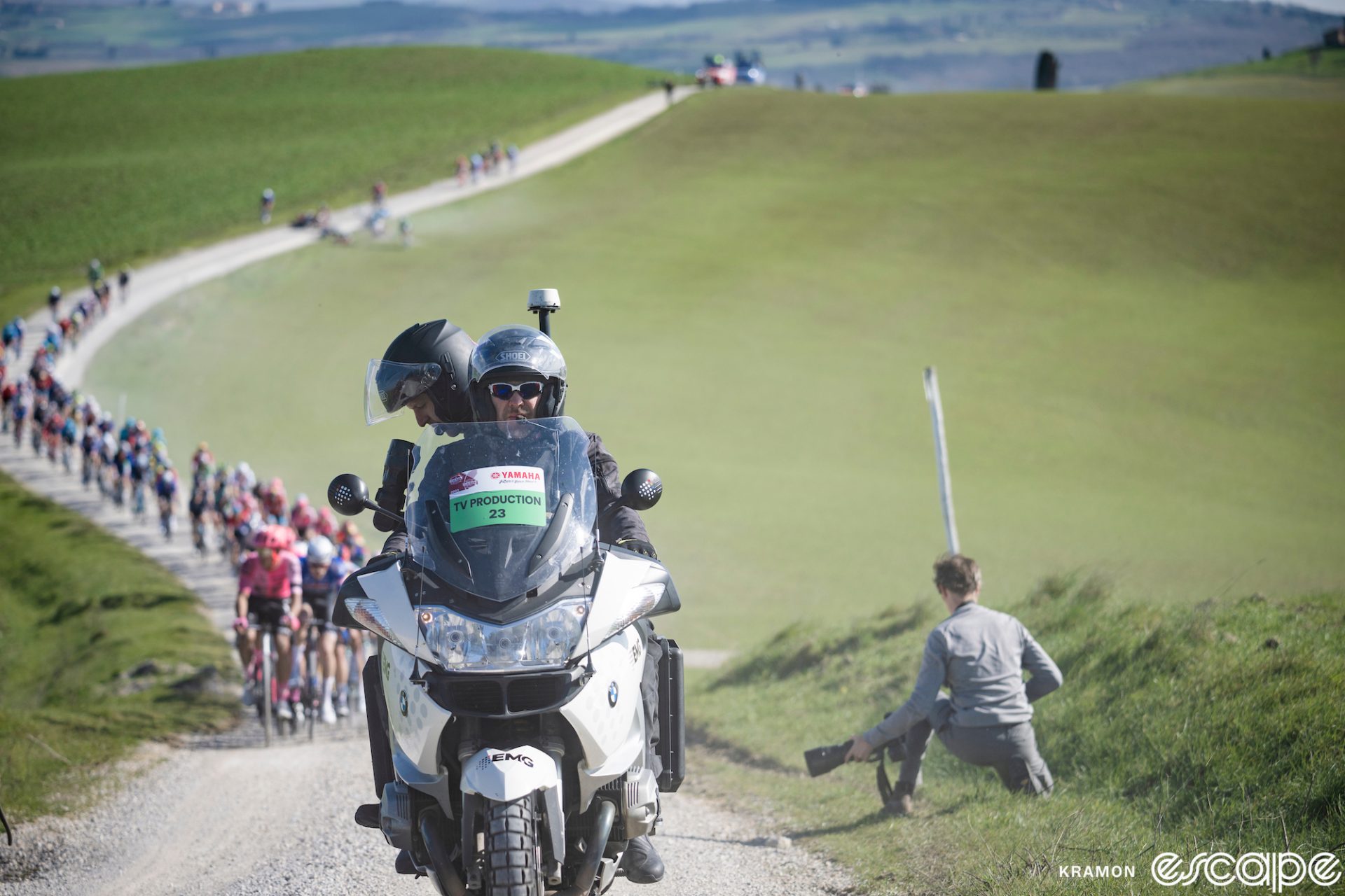 A television camera moto leads the pack at the 2023 Strade Bianche. The cameraman is peering through his eyepiece with the camera aimed back at the pack, which is strung out behind on a winding white gravel road that rolls through green fields.