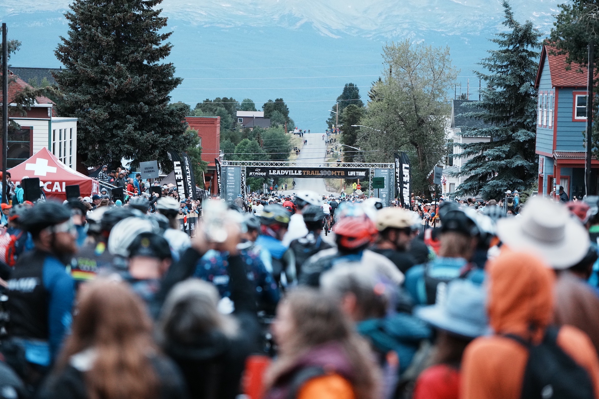 A view of the start of the Leadville Trail 100 mountain bike race, from a rider's perspective. The view looks down the street out of Leadville toward towering mountains in the background. In the foreground, a sea of riders is slightly out of focus as they await their start.