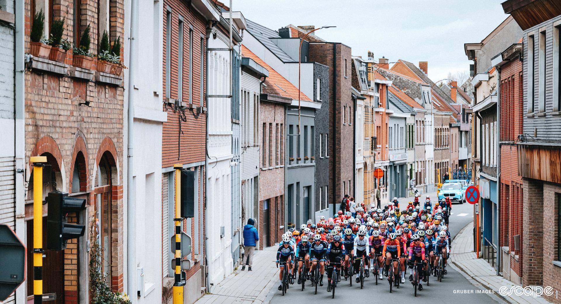 A peloton rides along, with buildings towering over them.