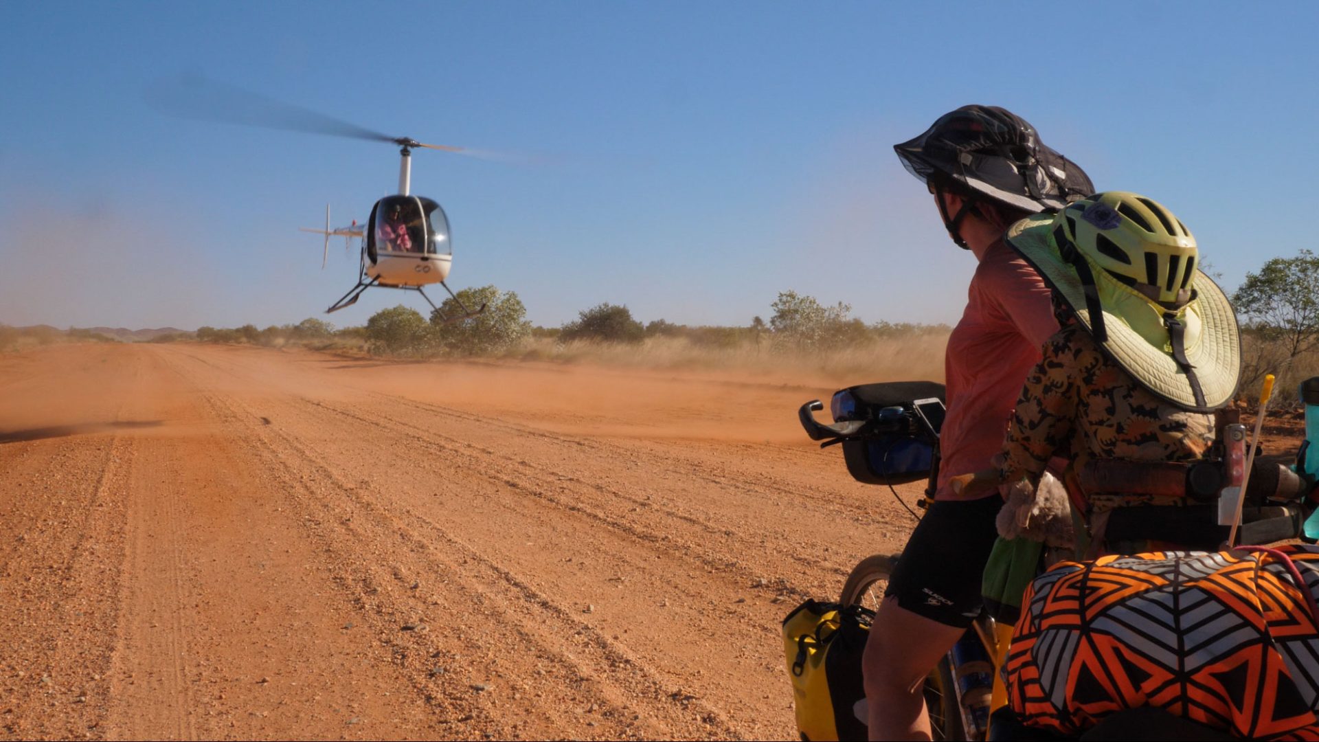 Wilfy and Nicola look at a small helicopter coming in to land on a red dirt road. 