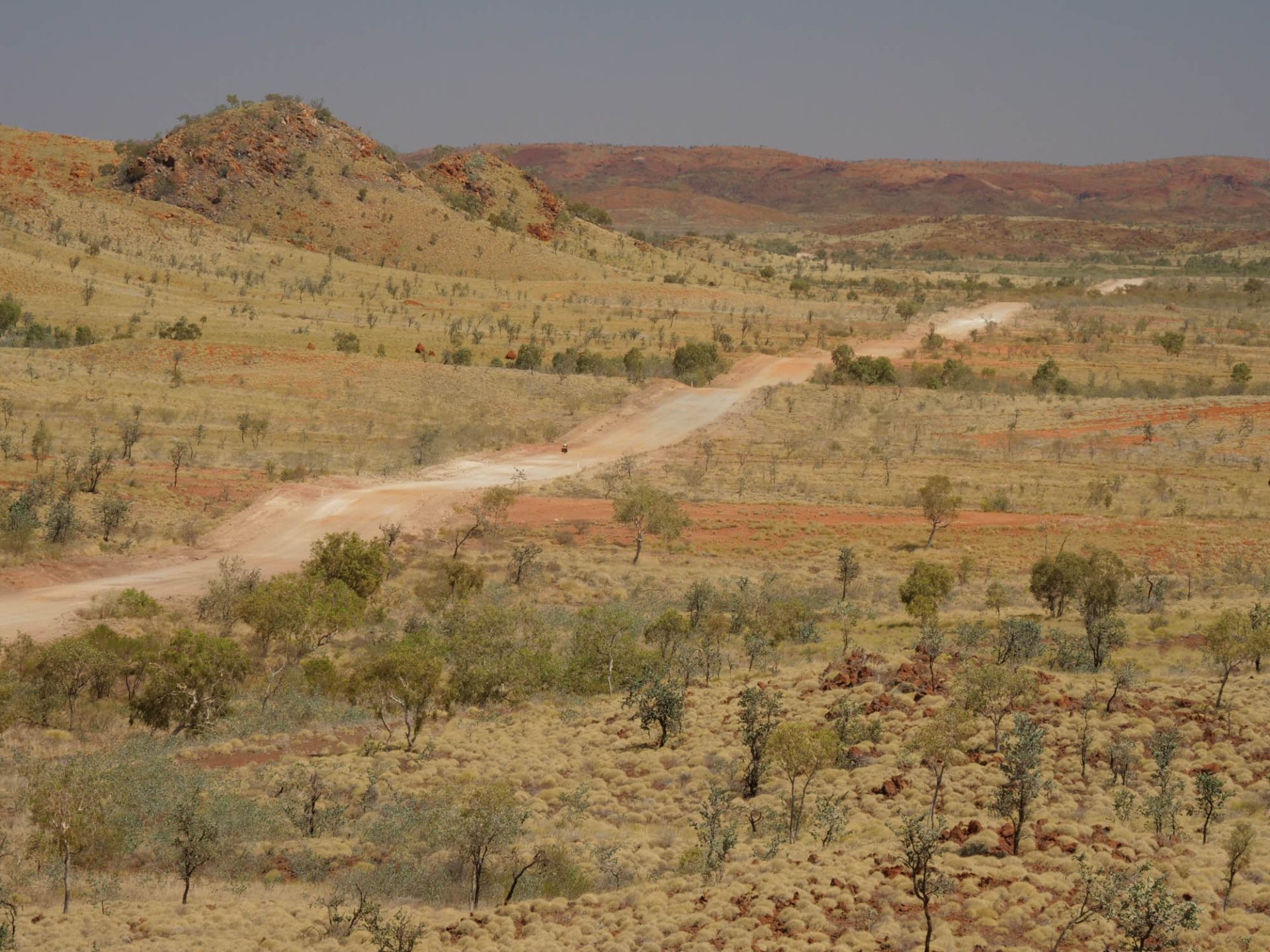 A sparse desert landscape with a rolling dirt road, with the tiny figure of a cyclist in black in the middle of frame. 