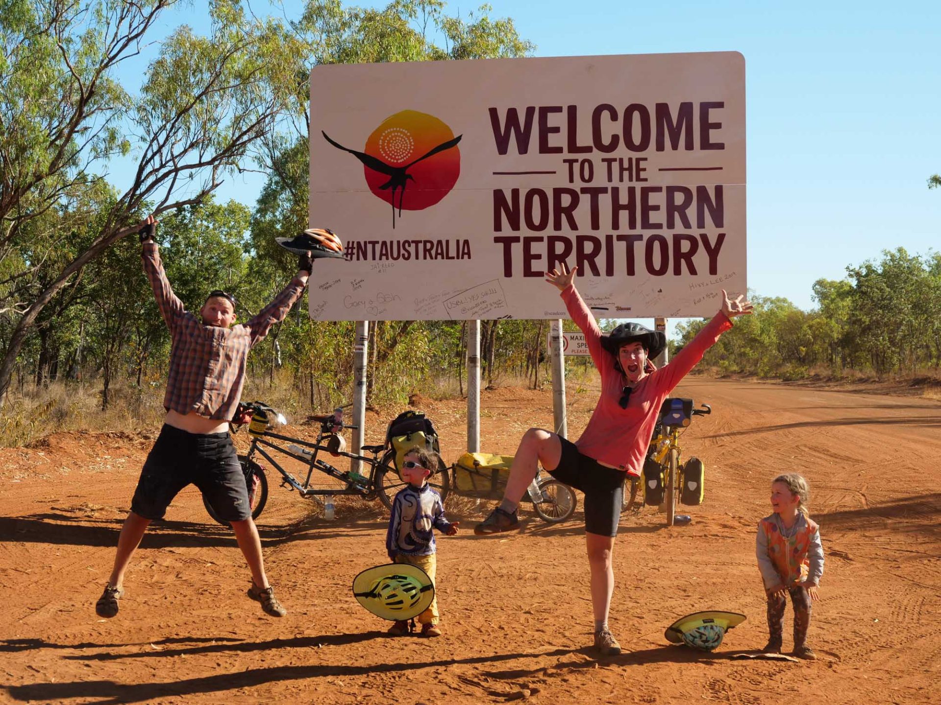 A sign in the background reads 'welcome to the Northern Territory'. All four members of the family pose in front of it. 