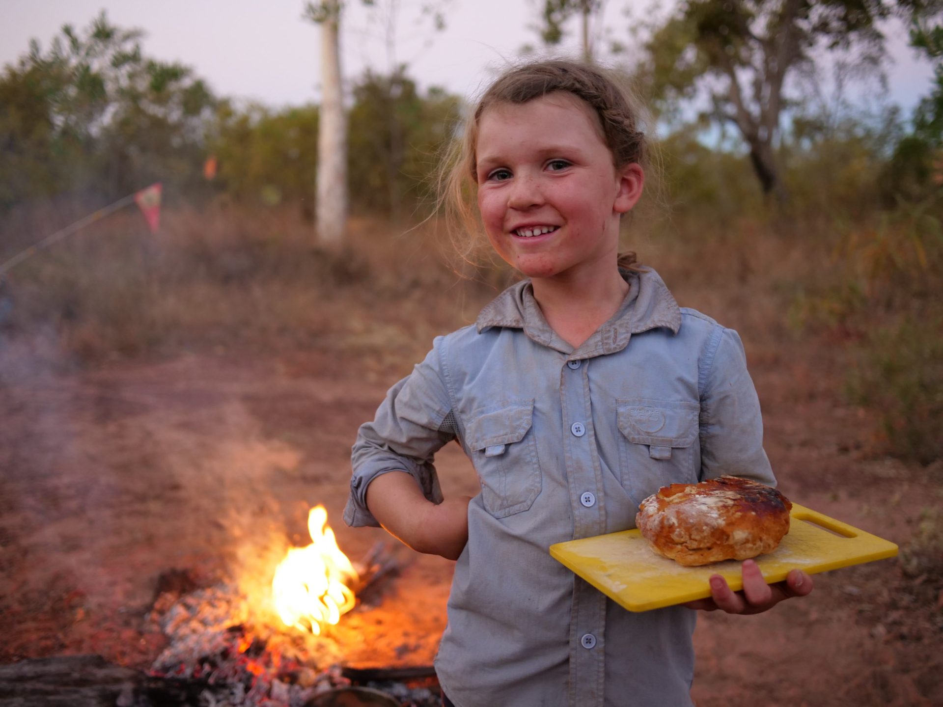 Hope poses, smiling, one hand on hip, next to a camp fire with some food. 