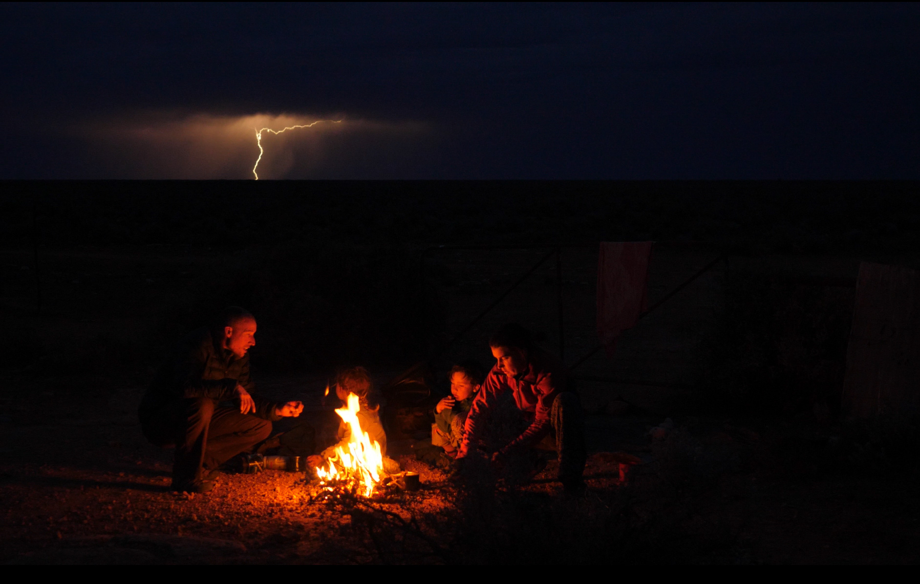 The family sits in a dark nothingness, lit only by a campfire. In the background is a storm with a dramatic lightning bolt piercing the sky. 