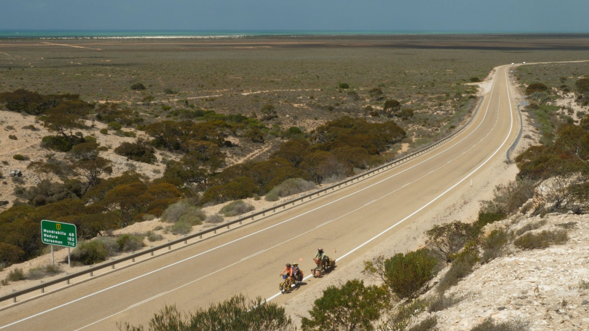Drone or altitude shot of the family touring along a long paved road, sparse landscape around them and the sea glinting distantly in the top of frame. 