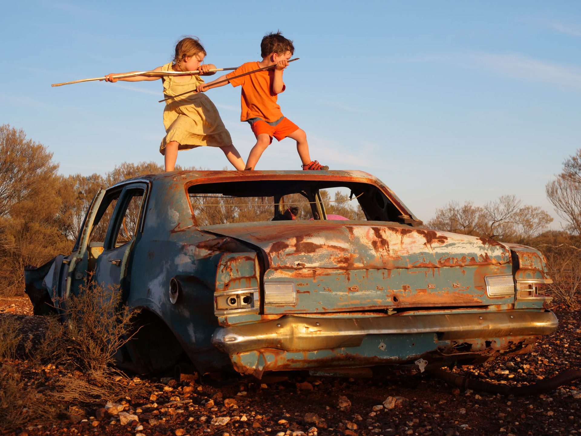 Hope and Wilfy, wielding sticks, stand in a ninja-like pose on the roof of an abandoned rusted out old car. 