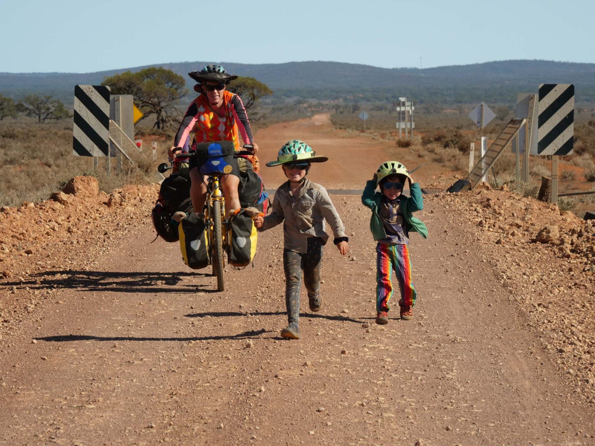 Nicola rides along a dirt road toward the photographer, both children running, smiling, alongside her. 