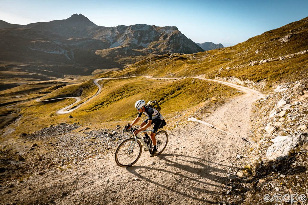 Mike Cotty rides up a gravel road in the Swiss Alps. He's shown all alone as the road switchbacks down into the valley below, amid treeless slopes of tan-brown grass and shrubs.