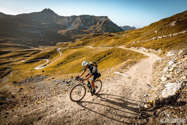 Mike Cotty rides up a gravel road in the Swiss Alps. He's shown all alone as the road switchbacks down into the valley below, amid treeless slopes of tan-brown grass and shrubs.