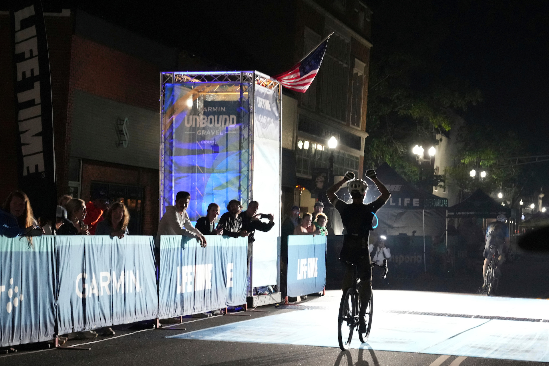 A lone finisher crosses the line at Unbound Gravel. It's dark, with the only light from floodlights on the line, and he's just behind another rider, and he is sitting up, raising his hands in a victory salute at having finished. A few people lean over the barriers to cheer.