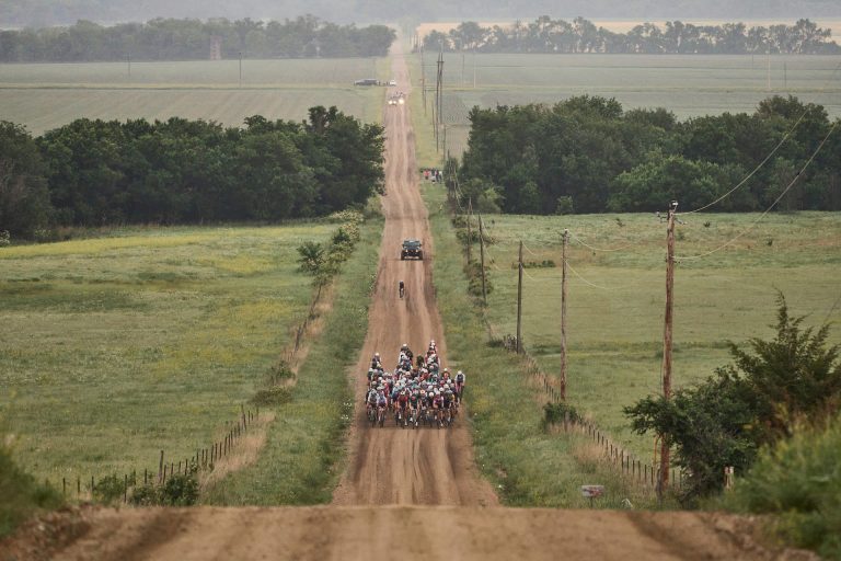 A pack of riders travels down a dirt road. The road is straight and disappears into the distance over rolling green hills. The tightly bunched pack of maybe 75 riders is isolated in the vast landscape of green fields and forest groves, with only a few follow cars behind.