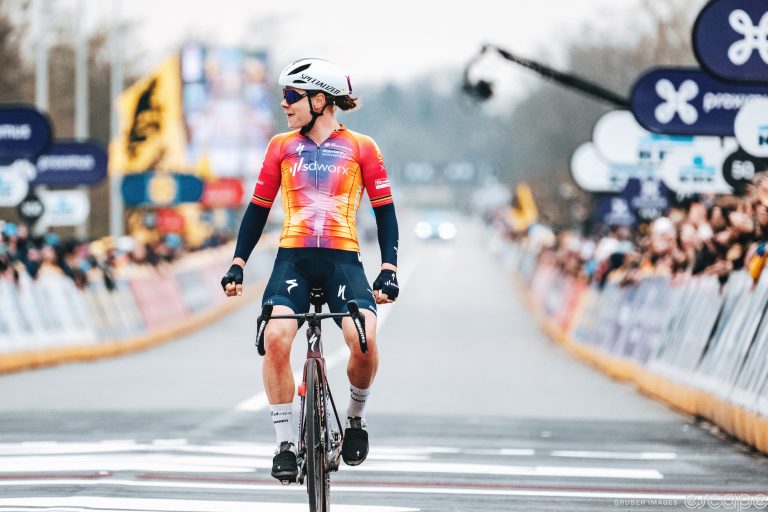 Lotte Kopecky sits up to celebrate her victory in the 2023 Tour of Flanders. She's looking to her right with an expression of confidence and satisfaction, and the road behind her is empty of riders as she crosses the line solo.