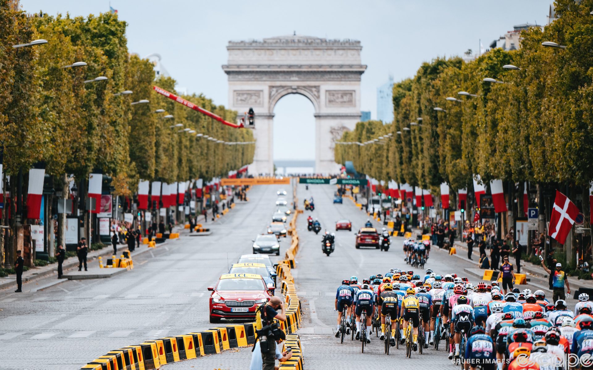 The pack races down the Champs-Elysées at the 2023 Tour de France. The road opens up in front of them, rising to the Arc de Triomphe.