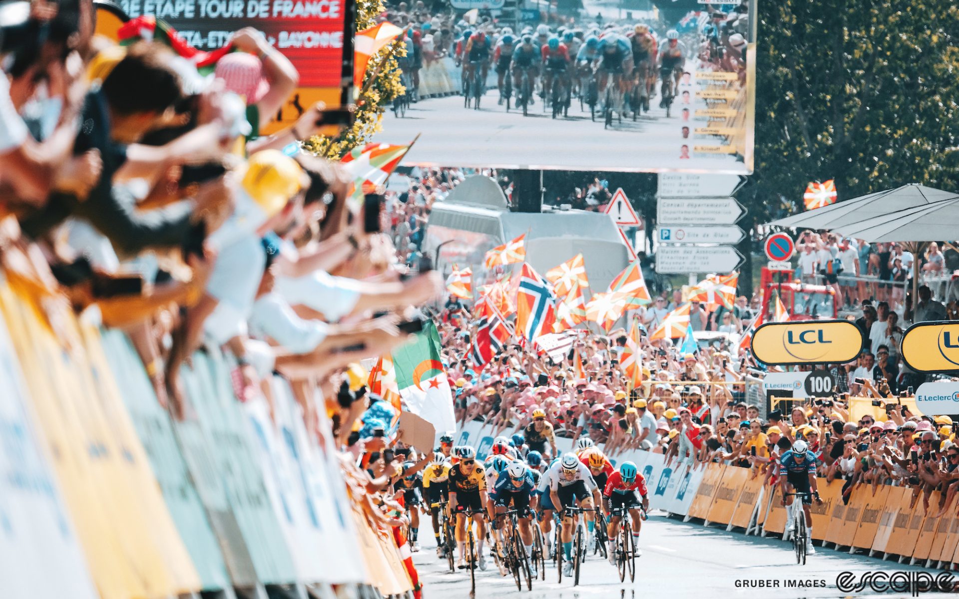 The Tour de France peloton sprints up a shallow rise at the end of the 2023 stage to Bayonne. Fans lean over the barriers and large colorful flags wave behind. Jasper Philipsen and Phil Bauhaus are seen at the front, while off to the right is Philipsen's teammate, Mathieu van der Poel, who has swung off from his leadout.