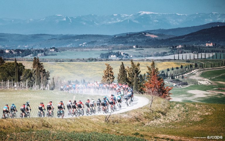 A peloton of cyclists races over dirt roads with snow capped mountains in the background.