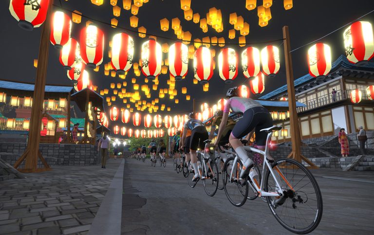 Zwift avatars race down an urban streetscape under rows of brightly lit paper lanterns.