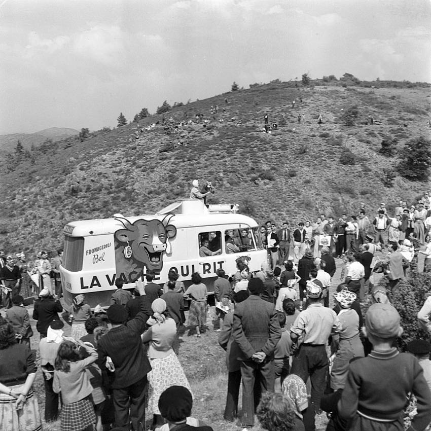 An old black and white picture of La Vache Qui Rit's van driving up a mountainside, surrounded by spectators. 