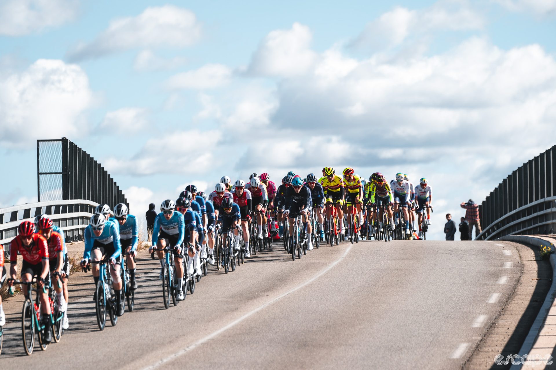 The pack crosses an overpass in the Tour de la Provence. The group is finally riding on dry roads under the sun but is strung out from the pace and crosswinds.