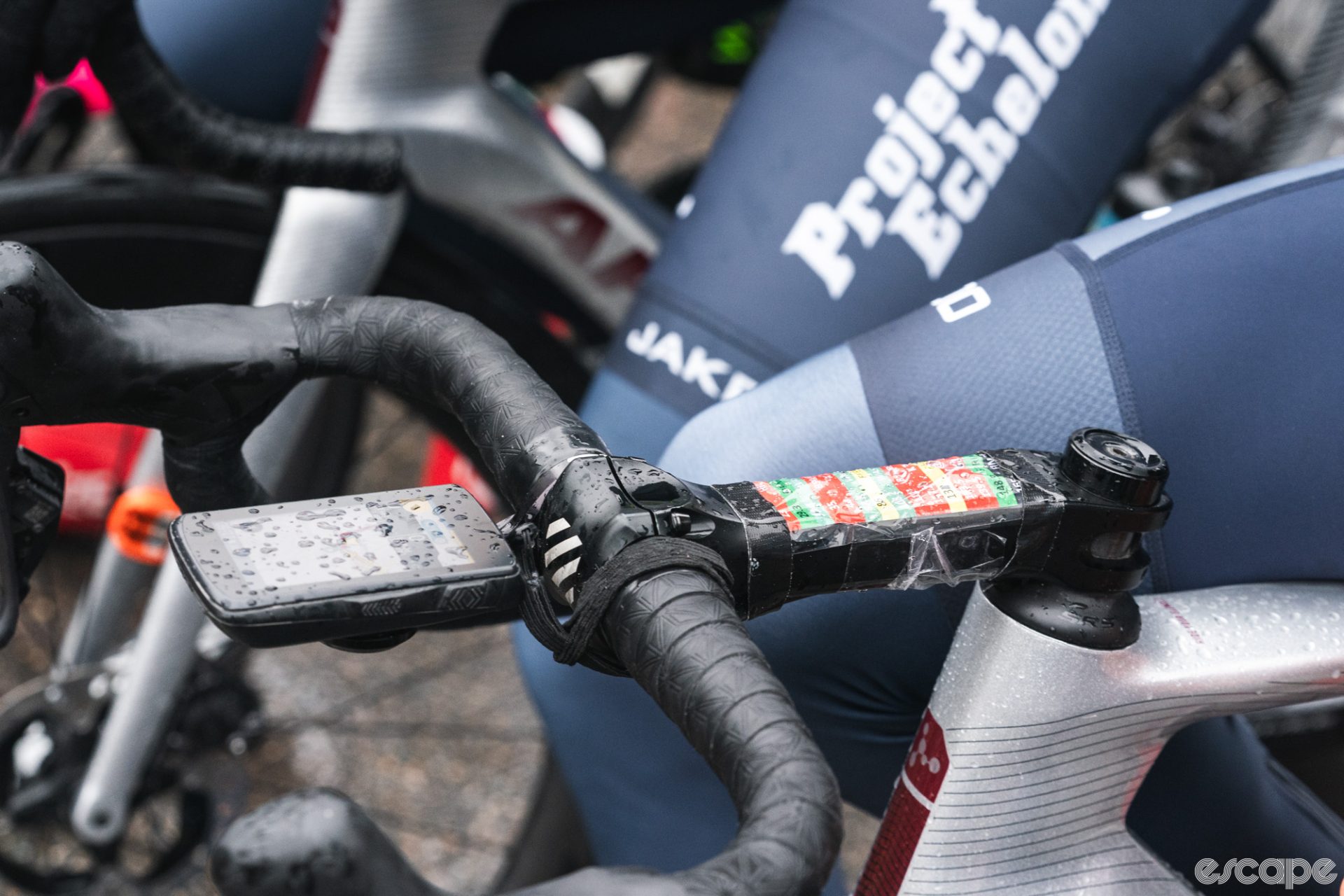 A Project Echelon Argon-18 team bike sits at a race start. The head unit is dotted with rain, and the route profile is taped to the stem and already getting wet, colors bleeding slightly.