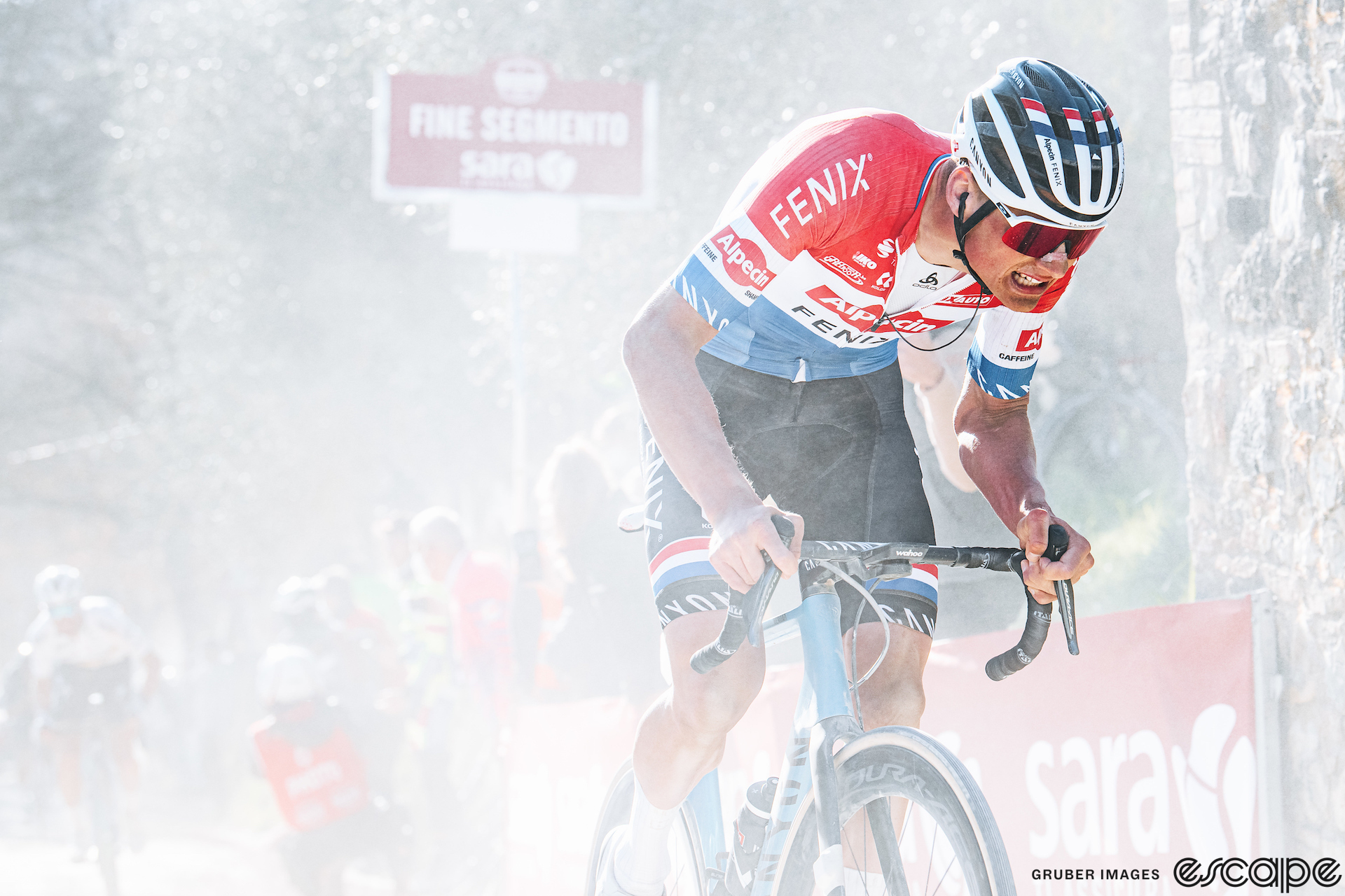 Mathieu van der Poel grimaces as he attacks out of the saddle on a late gravel sector in Strade Bianche. He's bent low over the bike, wearing the Dutch national champion's jersey in bands of red, white, and blue. White dust rises all around him.