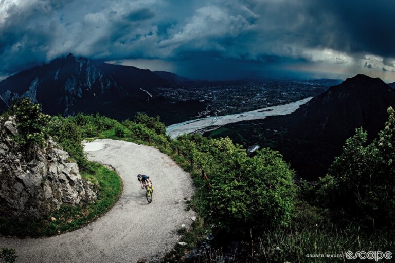 A lone rider rounds a switchback on a climb amid a lush landscape. Behind, a storm looms over the mountains and lake in the valley below.