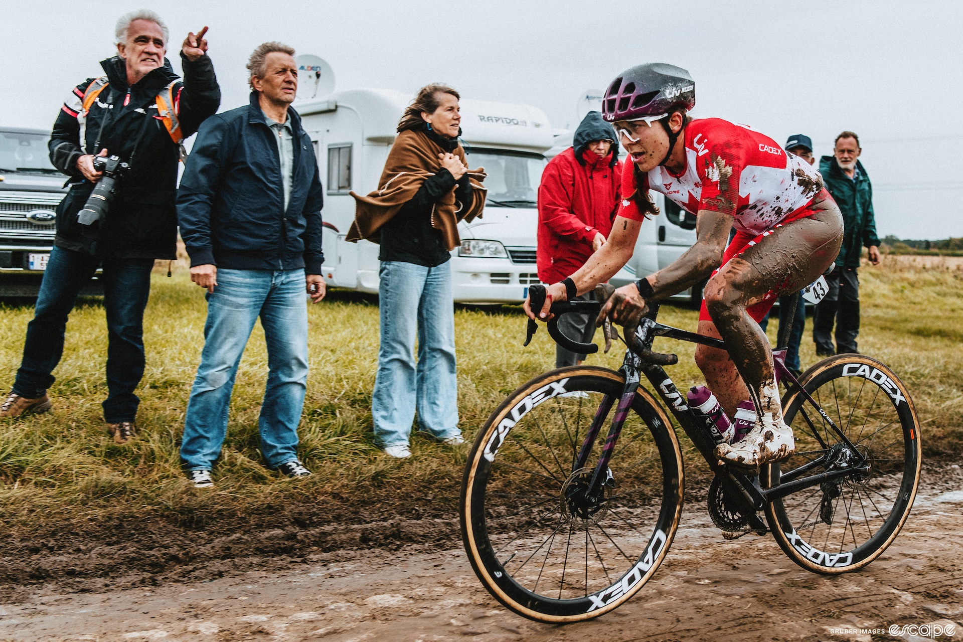 Alison Jackson rides across cobbles at the 2021 Paris-Roubaix, covered in mud. She has clearly fallen down at some point but is back on her bike looking grumpy. 