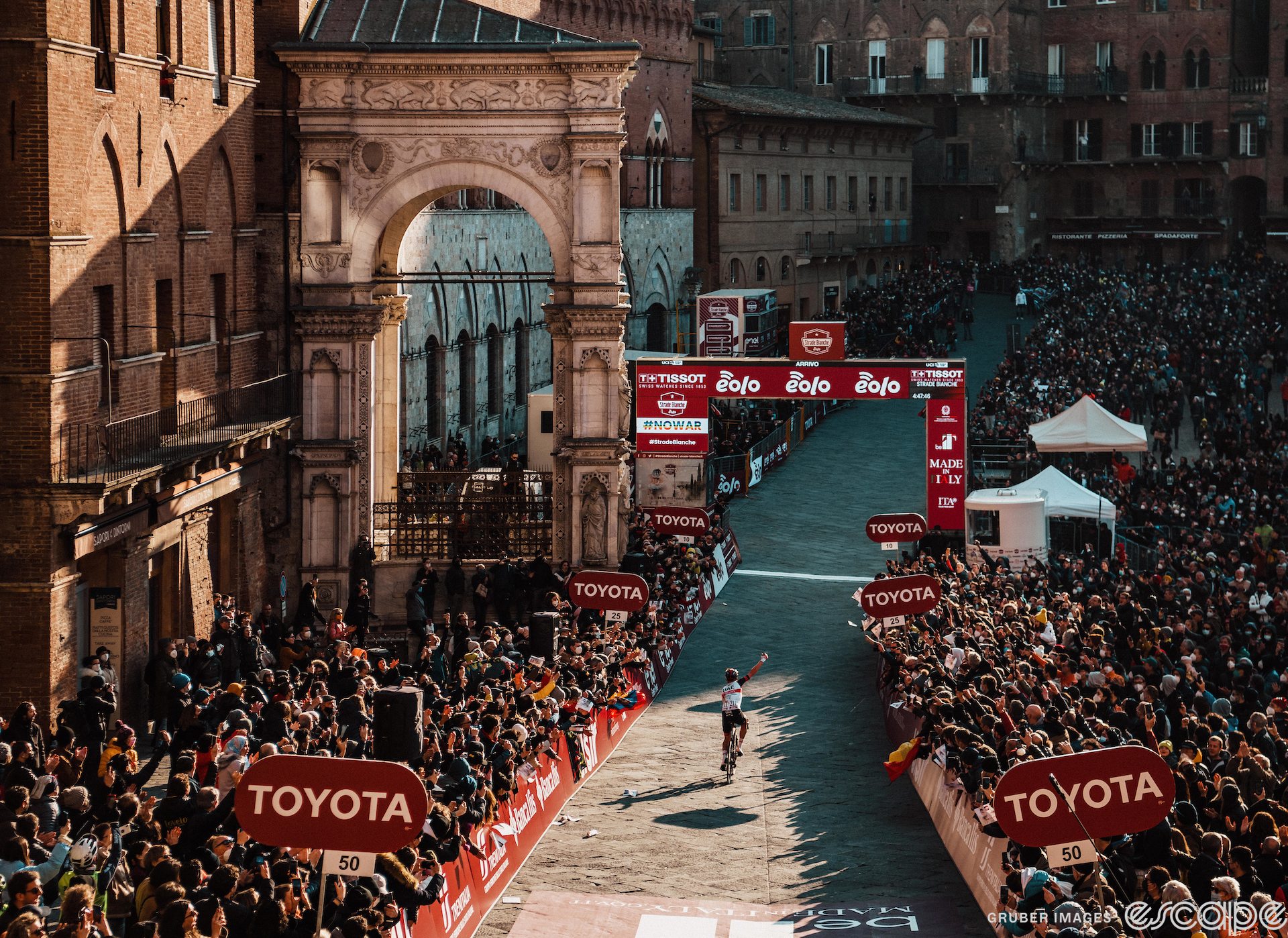Tadej Pogačar approaches the finish line of the 2022 Strade Bianche. The wide shot takes in the mass of crowds in Siena's iconic Piazza del Campo, with red-brick medieval buildings crowded in close next to a large arch.