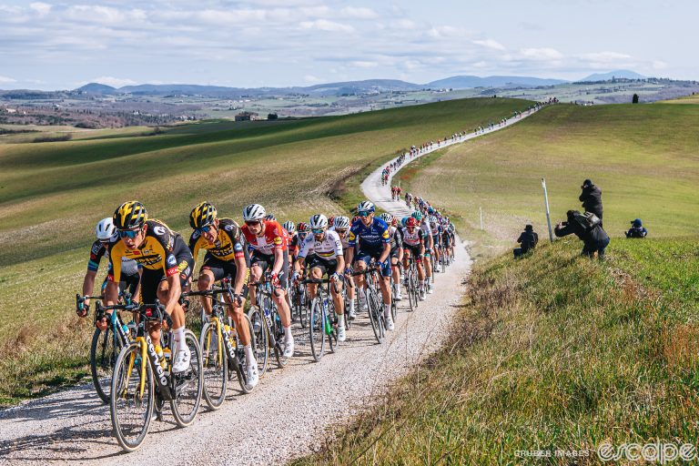 Wout van Aert leads a line of riders along a white gravel road at Strade Bianche. The road twists away lazily in the background through greening fields and Tuscan hills.